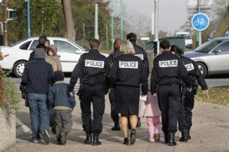Un groupe de six policiers escorte une famille de cinq étrangers. Entre leurs jambes, on aperçoit une fillette qui doit avoir quatre ans, toute habillée de rose.
